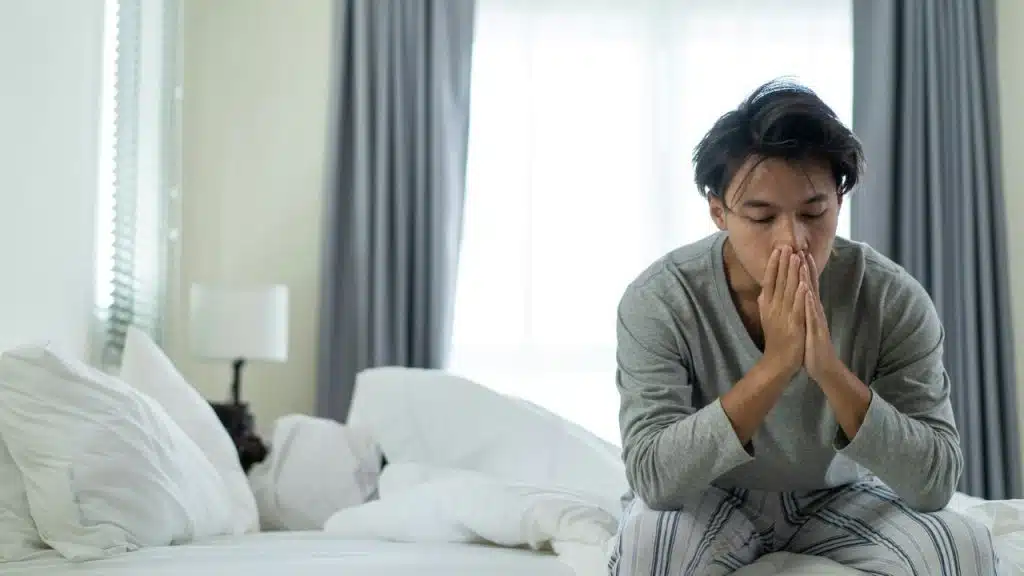A man sits on the edge of a bed with white linens, holding his hands to his face.