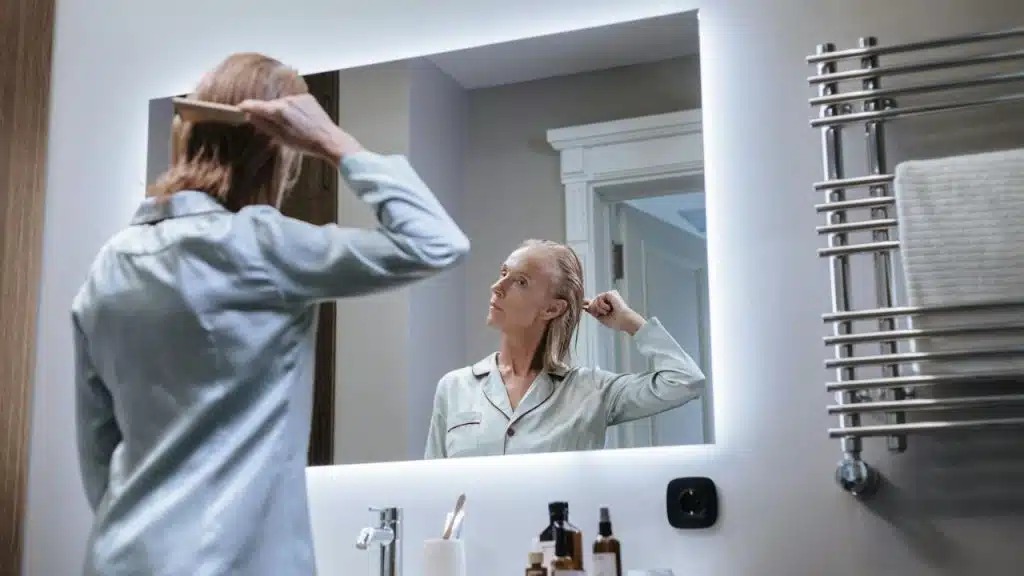 A woman in light blue pajamas combs her wet hair while looking into a lighted mirror.