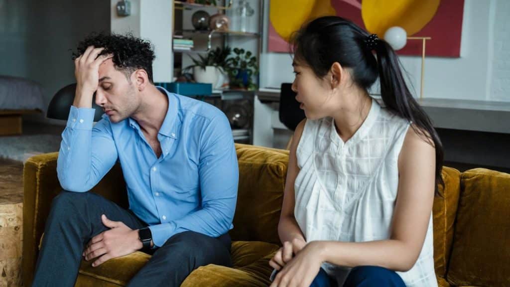 A man sits on a yellow sofa with his hand on his forehead beside a woman.