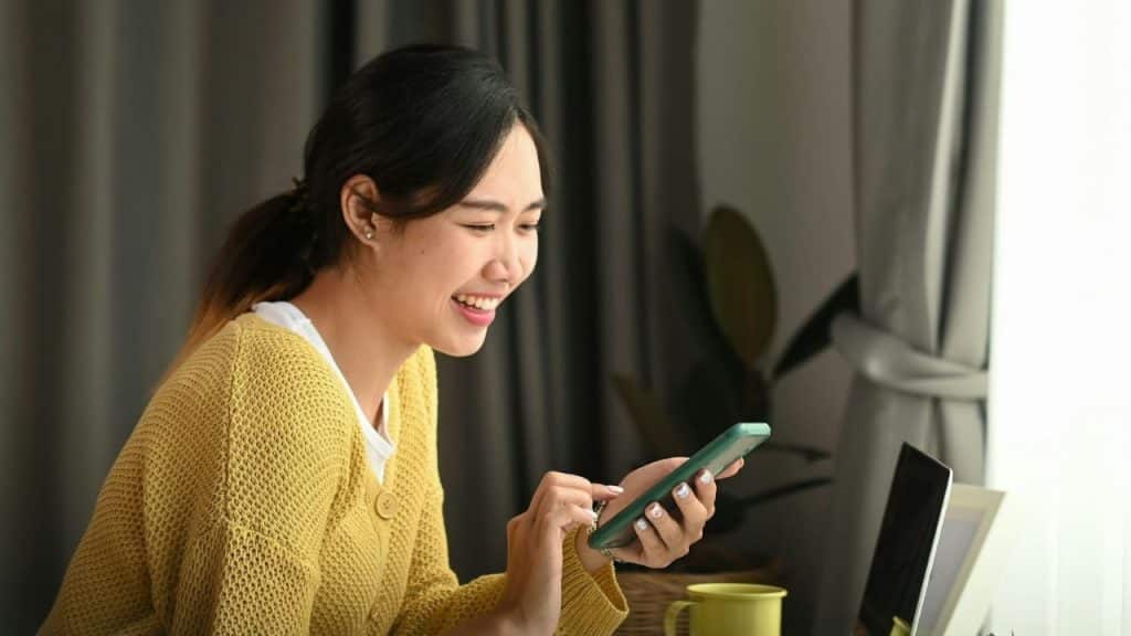 A woman in a yellow cardigan smiles while looking at a smartphone near a window.