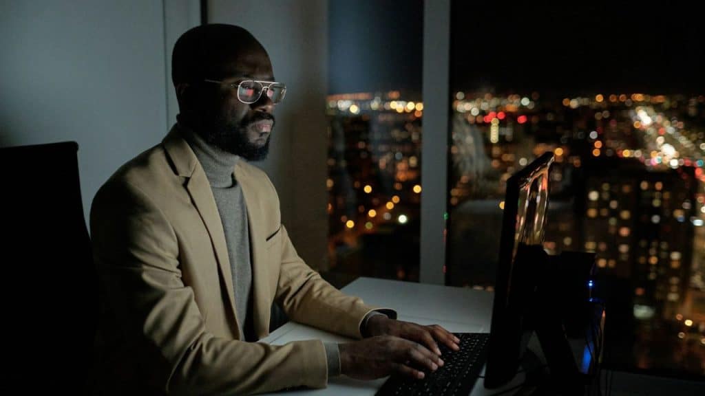A man wearing glasses and a blazer works on a computer at night overlooking a city.