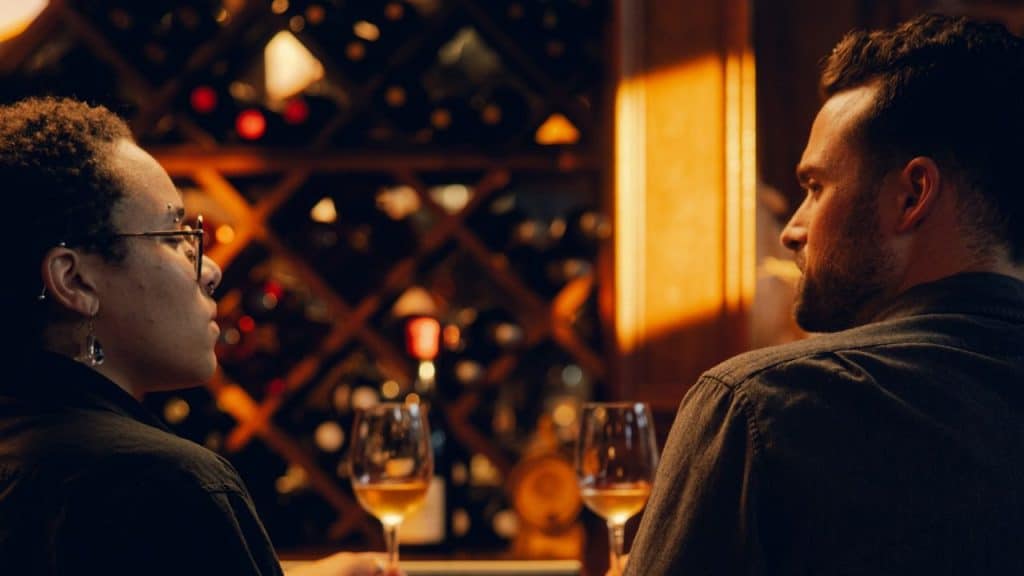 Two people sit at a bar with wine glasses, facing a wall of wine bottles.