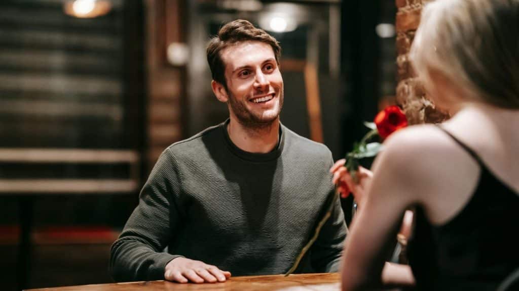 A smiling man sits at a wooden table looking at a woman holding a rose.