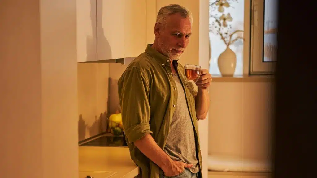 A gray-haired man in a green shirt leans against a kitchen counter holding a clear mug.