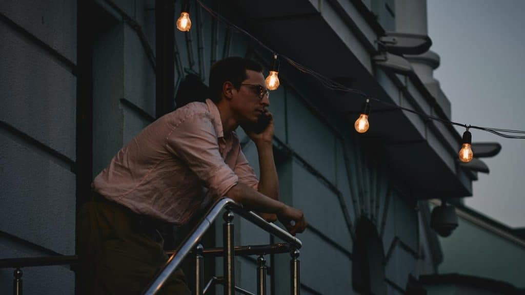A man leaning on a balcony railing while talking on a phone at dusk.