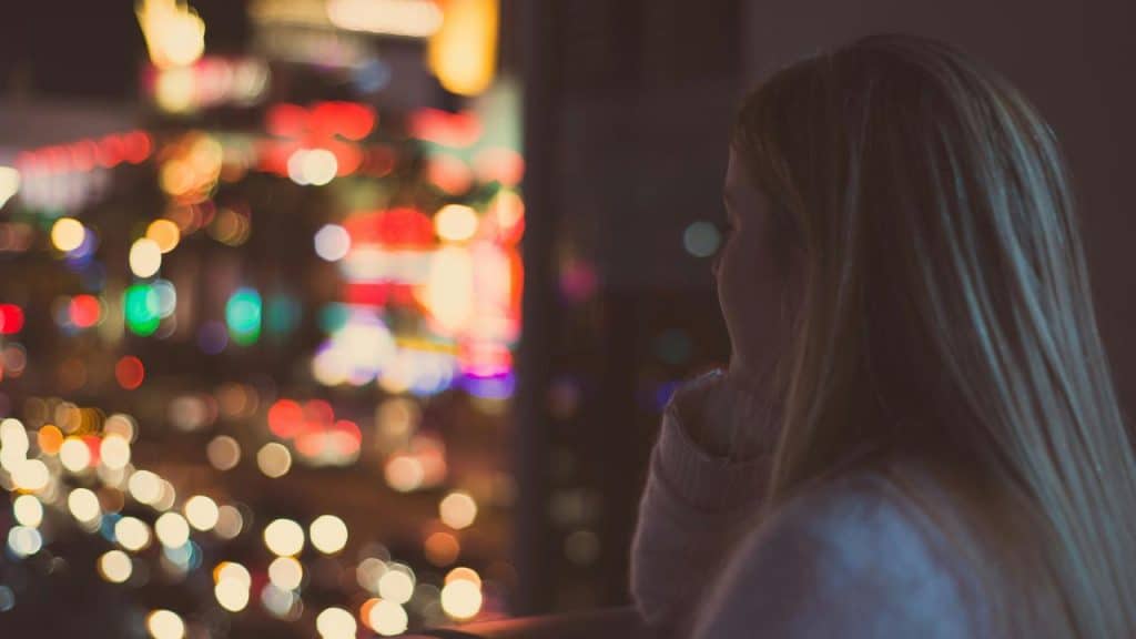 A woman looking out over colorful city lights at night from a balcony.