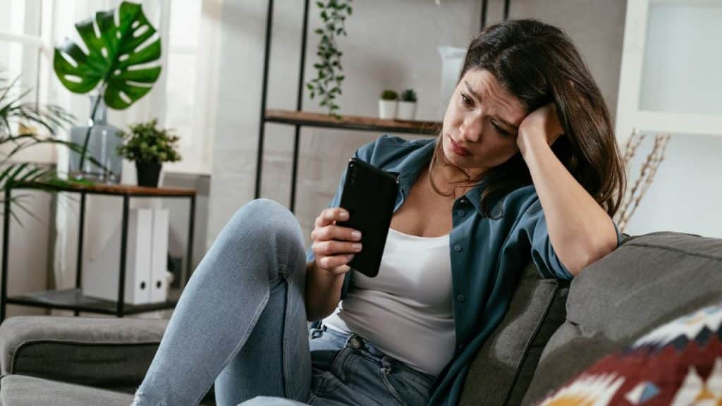 A woman sits on a couch looking at a smartphone with a concerned facial expression.
