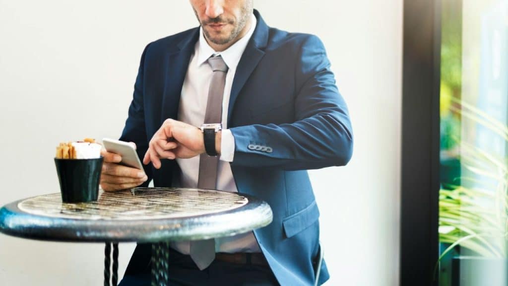A man in a suit sits at a table checking his wristwatch while holding a smartphone.