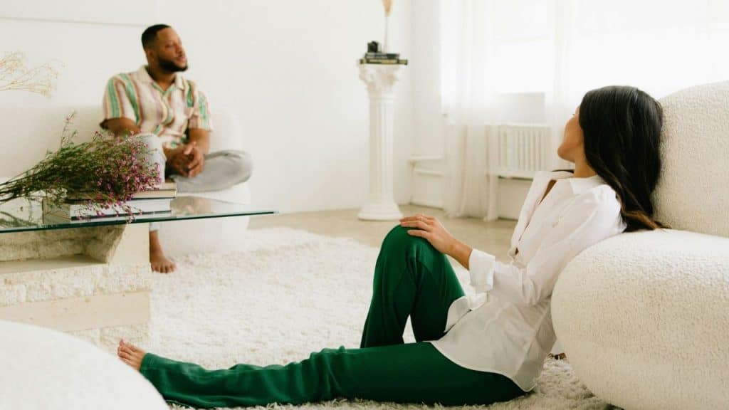 A man sits on a white sofa while a woman sits on the floor nearby.