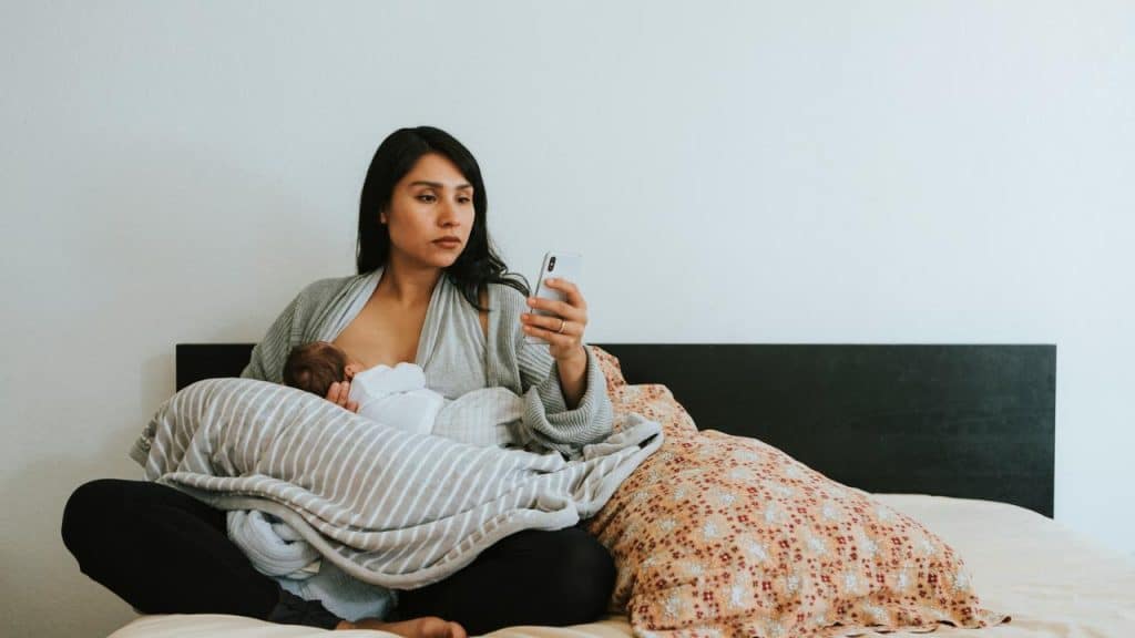 A woman sitting on a bed holding a baby while looking at her phone.