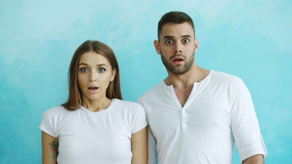 A man and a woman in white shirts stand against a blue wall with surprised expressions.