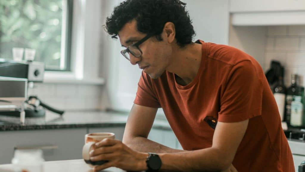 A man wearing glasses and an orange shirt stands in a kitchen holding a ceramic mug.
