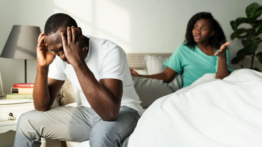 A man sits on a bed with his head in his hands while a woman speaks.