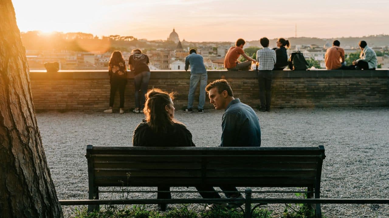 A man and woman sit on a park bench at sunset overlooking a city.