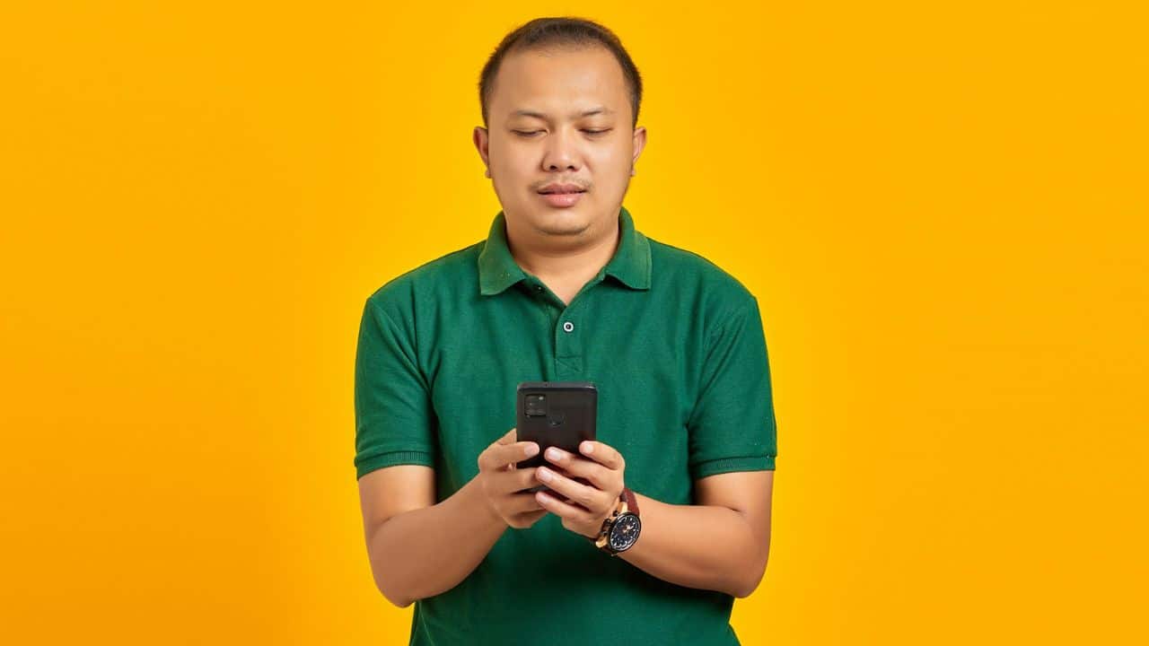 A man in a green polo shirt stands against a yellow background, looking at his smartphone.