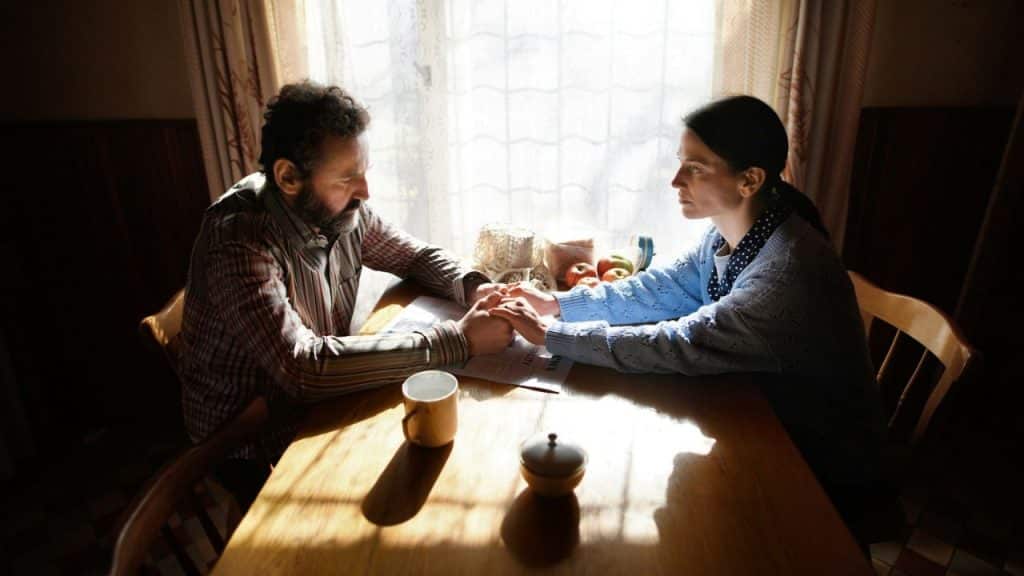 A man and woman sit at a wooden table, holding hands while looking at each other.