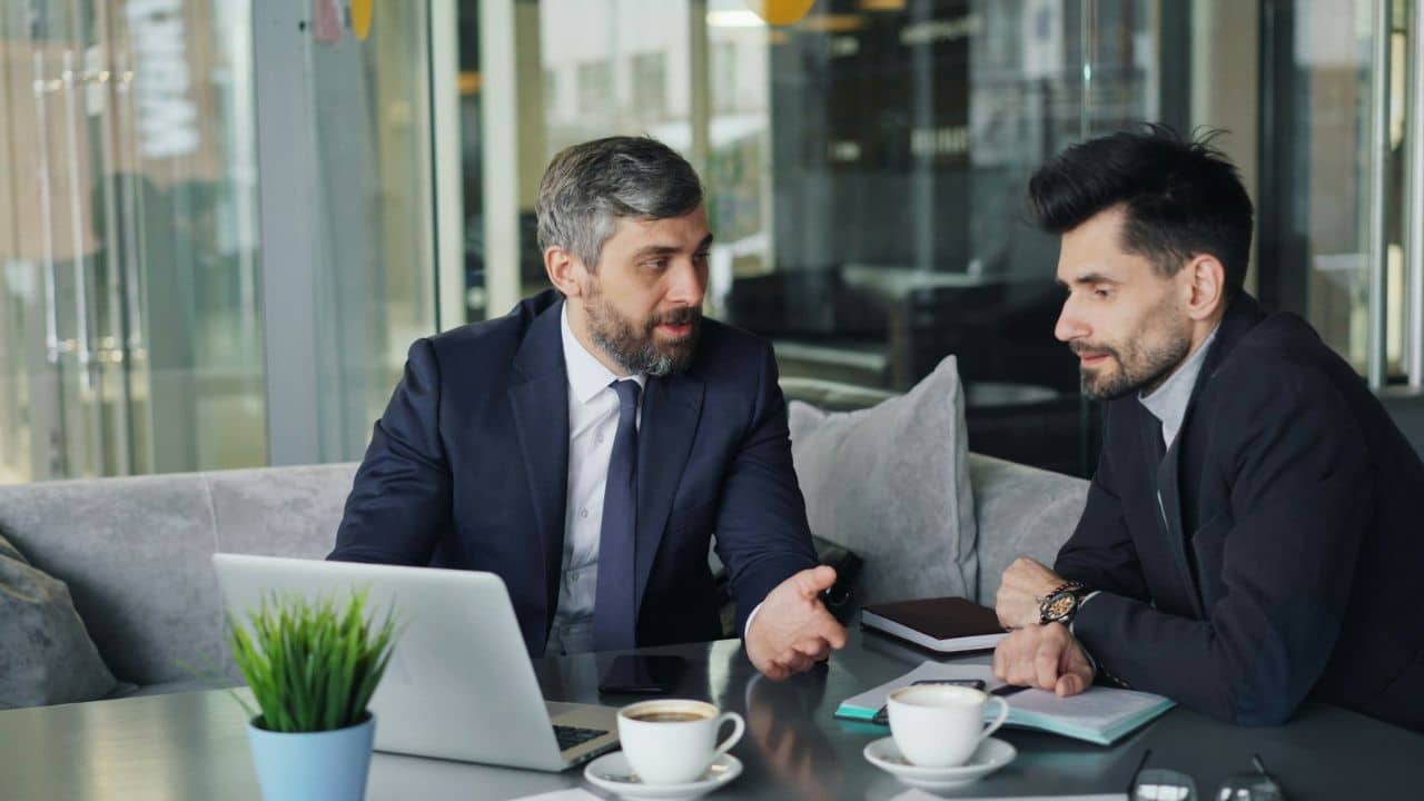 Two men in suits sit at a table with a laptop and coffee, talking.