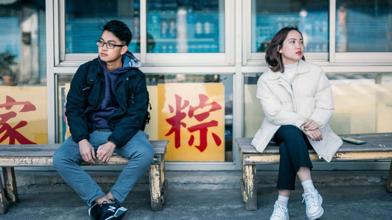 A man and woman sit apart on a wooden bench in front of a window.