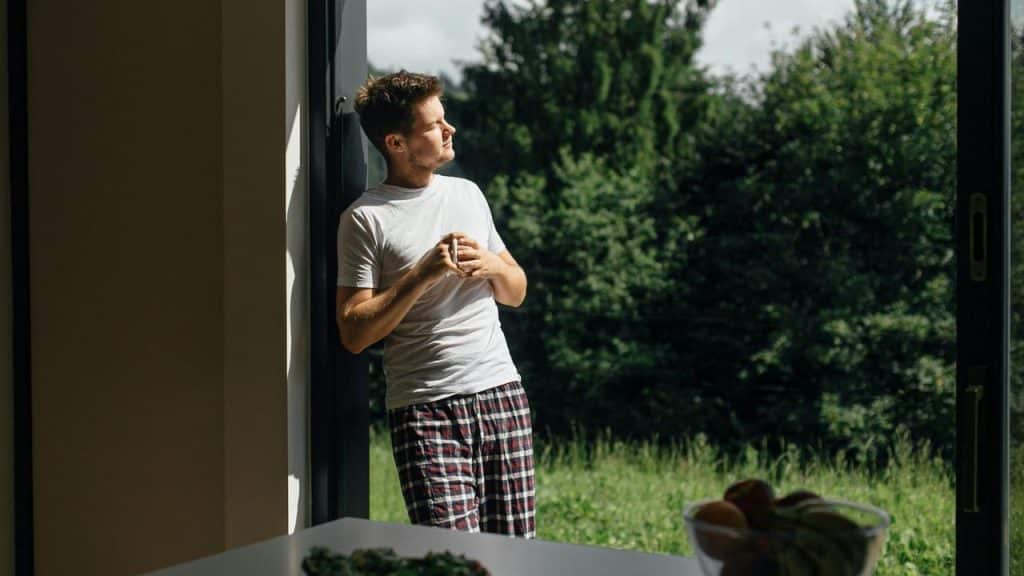 A man in pajamas holding a cup and looking outside through a large doorway.