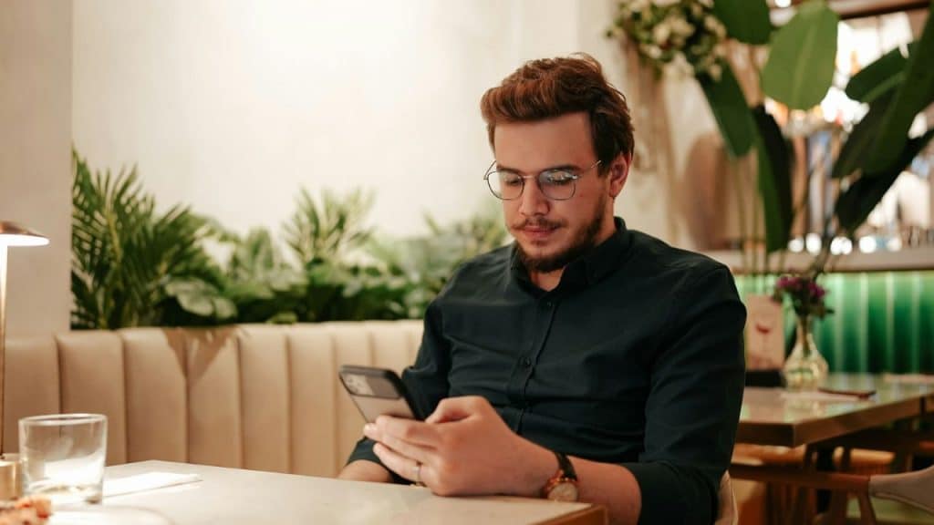 A man wearing glasses sits at a restaurant table and looks at his handheld smartphone.
