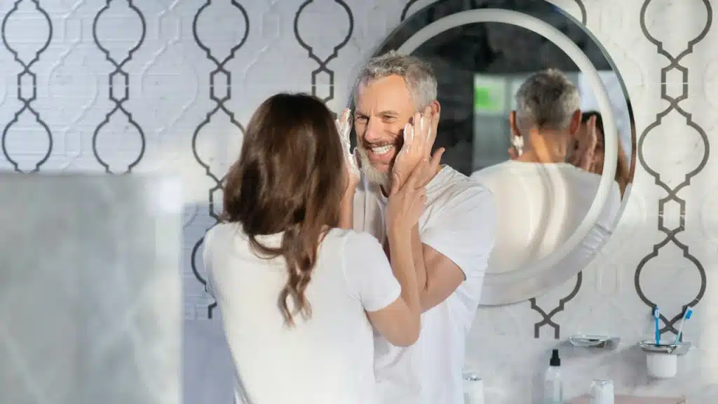 A woman holding a man’s hands as he smiles at her and tries to apply shaving foam to his beard as they stand in a bathroom.