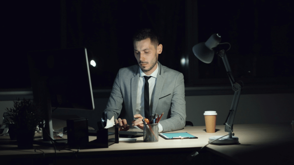 A man in a suit works at a computer in a dark office lit by a lamp.