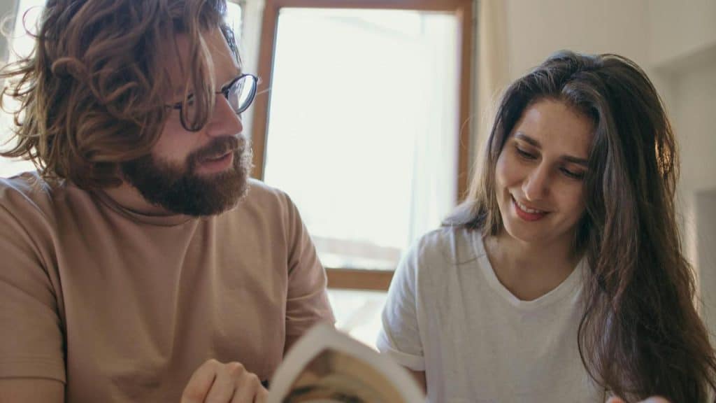 A bearded man and a woman sitting together and looking at an open book by a window.