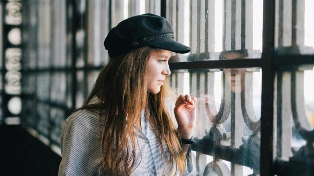 A woman wearing a dark cap gazing thoughtfully out a window with her reflection visible.