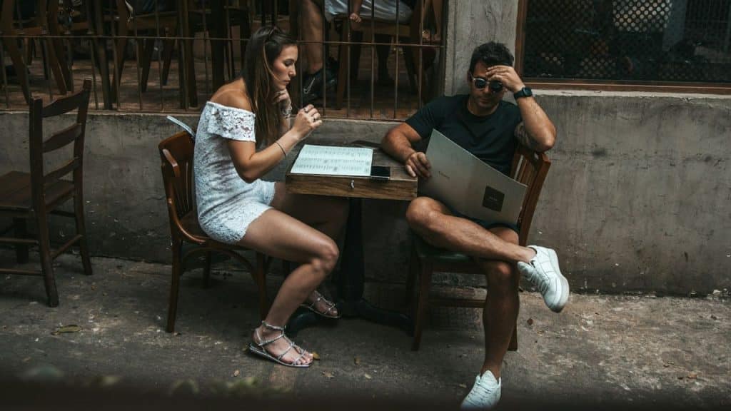 A man and woman sit at an outdoor cafe table looking down at handheld menus.