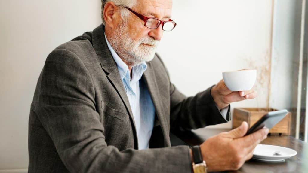 An older man in glasses holds a coffee cup while looking at a smartphone.