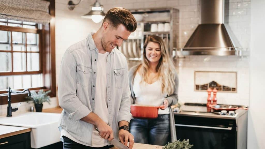 A man in a denim shirt chops vegetables while a woman carries a red pot.
