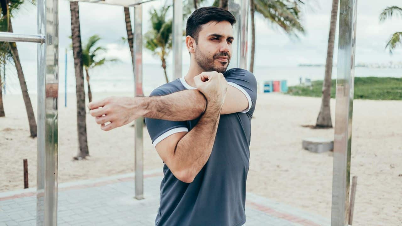A man in a grey shirt stretches his arm at an outdoor beach gym area.