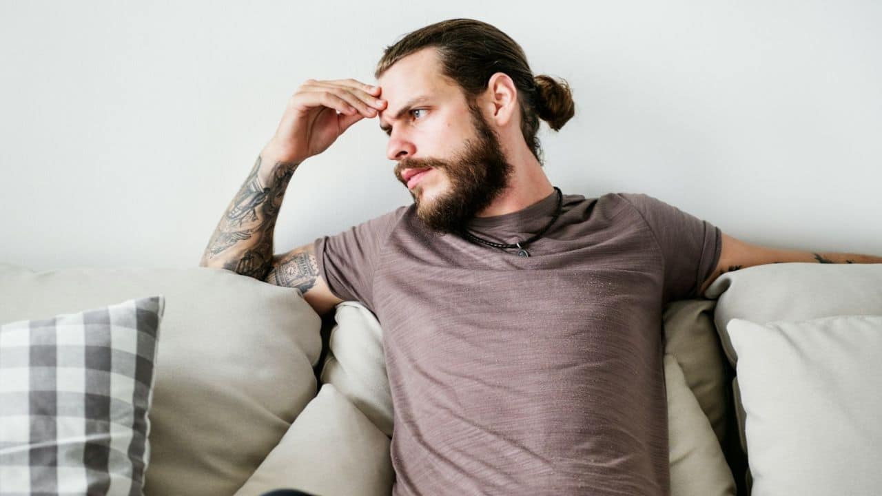 A man with a beard and tattoos sits on a couch while touching his forehead.