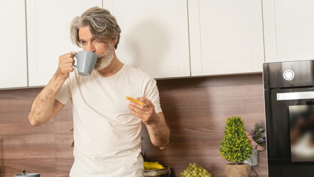 A man with gray hair drinks from a mug while holding a piece of toast.