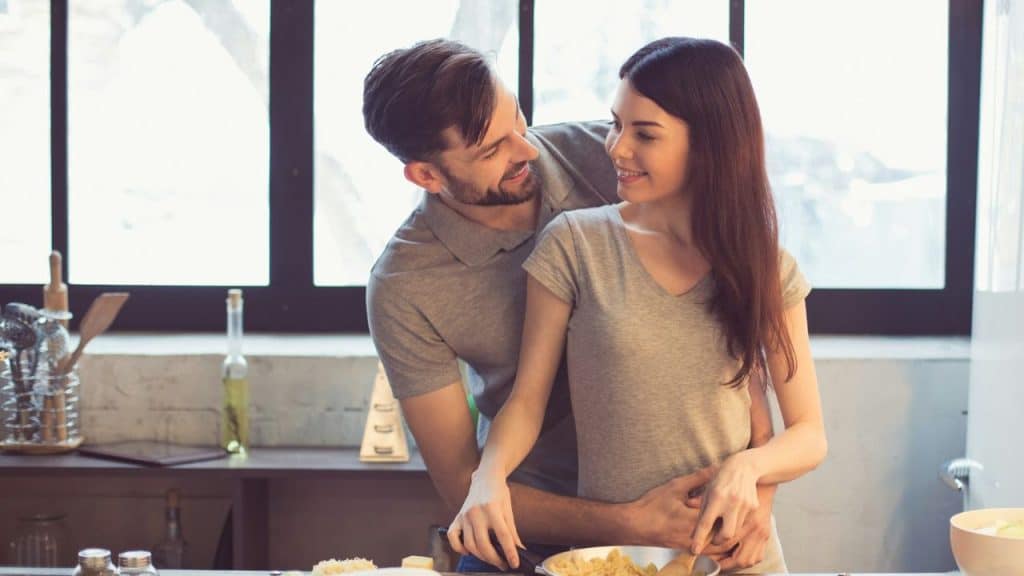 A couple smiles at each other while cooking together in a kitchen.