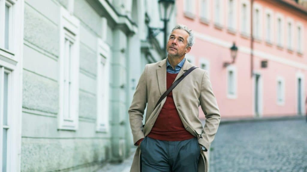 A man in a tan coat and red sweater walks down a European-style street.