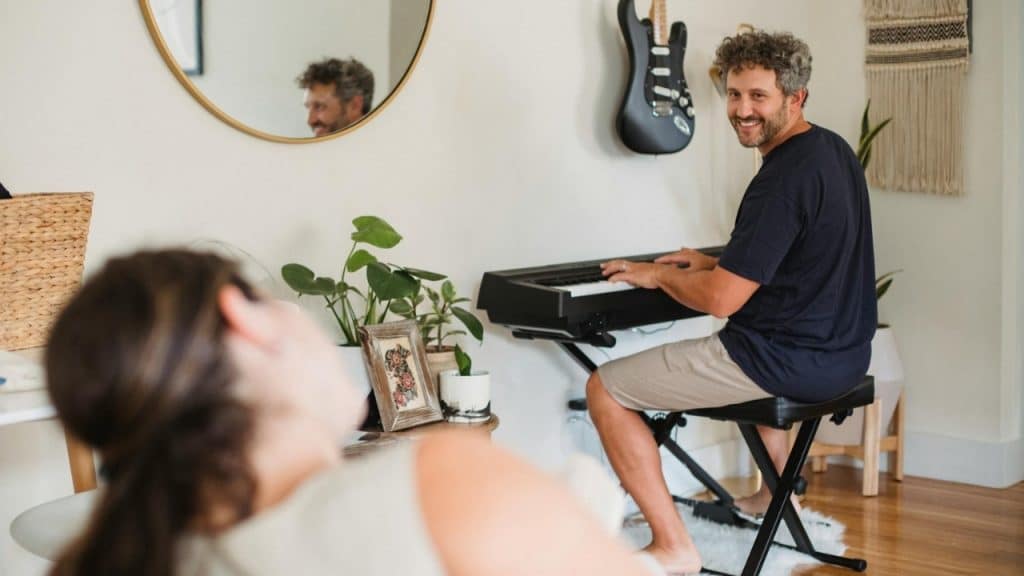 A smiling man plays a keyboard in a room while a woman's back is visible.