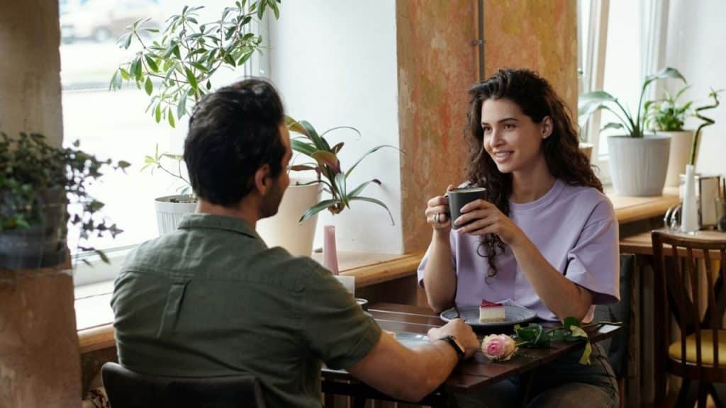 A man and woman sit at a cafe table with coffee, cake, and a rose.