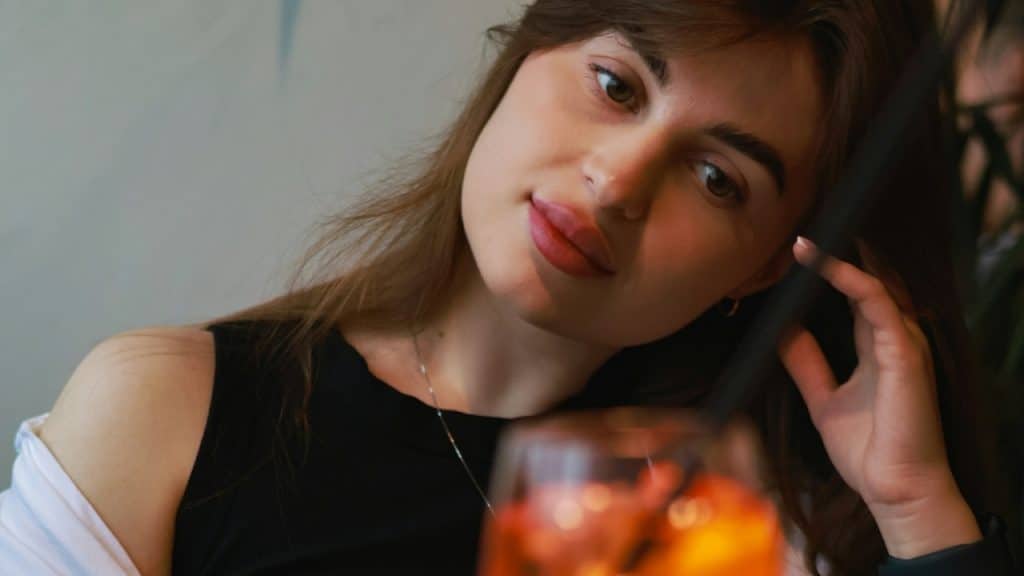 A woman leans her head on her hand and looks thoughtfully toward a blurred orange drink.