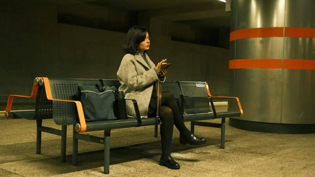 A woman in a grey coat sits on a metal bench using her phone at night.