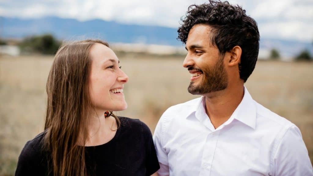 A smiling man and woman look at each other in an outdoor field with mountains.