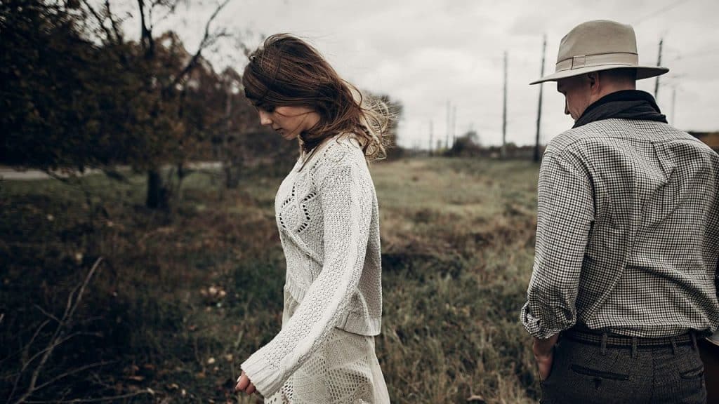 A woman walks through a field looking down, with a man following behind her.