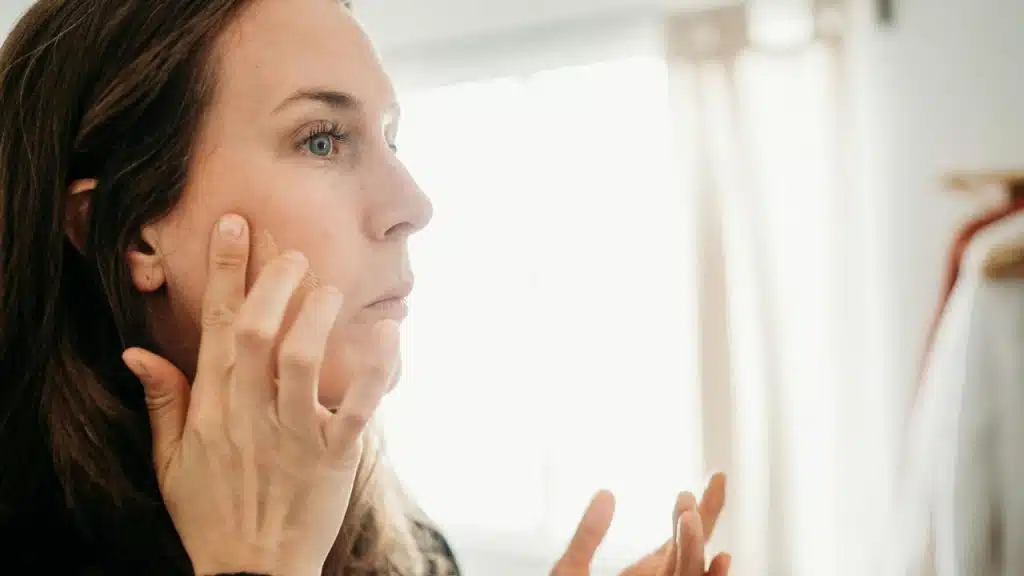A woman touching her face while examining her skin in a bright room.