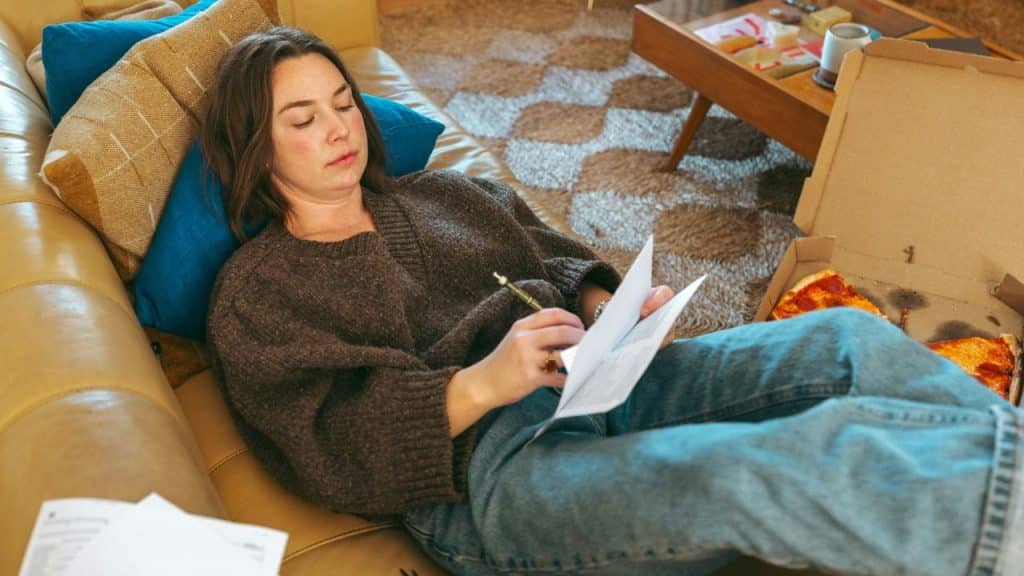 A woman reclines on a couch while writing on papers next to an open pizza box.
