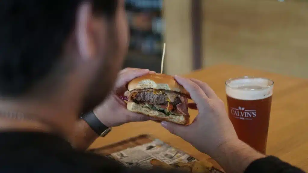 A person holds a large hamburger with both hands next to a glass of dark beer.