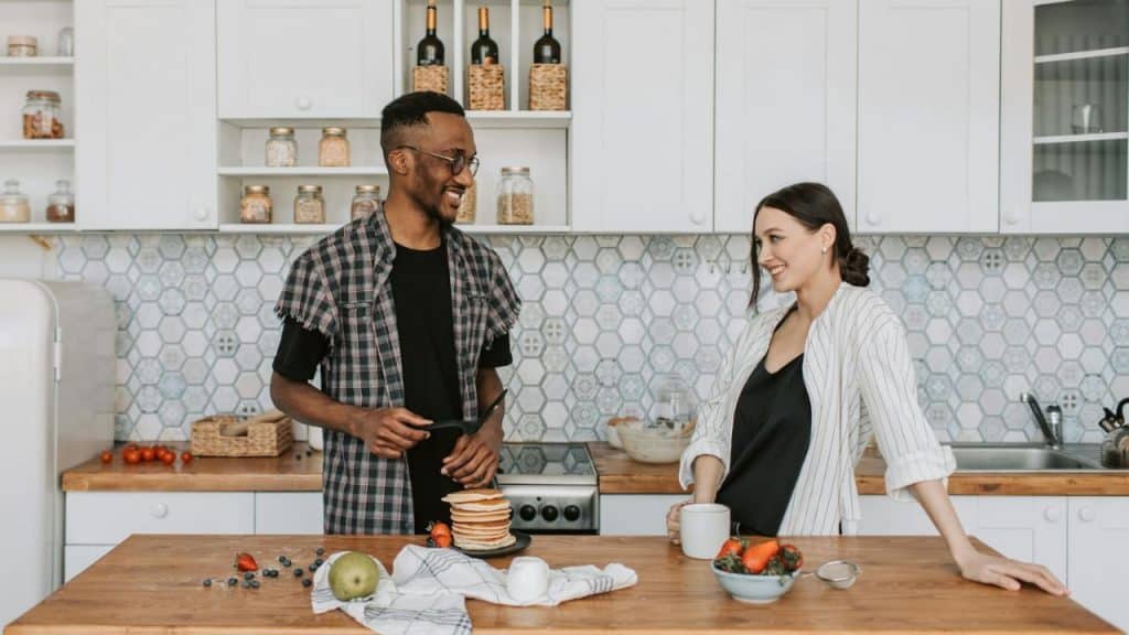 A man and woman smile at each other in a kitchen near a stack of pancakes.