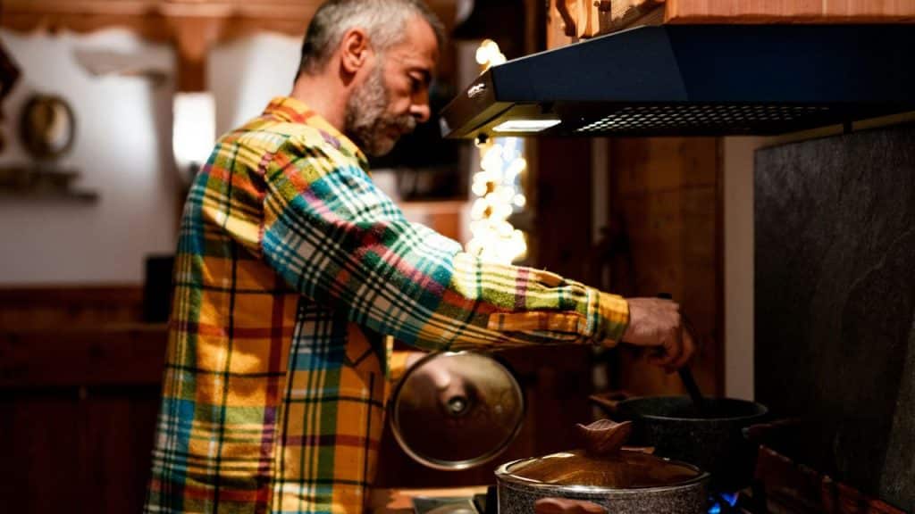 A man in a yellow plaid shirt stirs a pot on a stove in a kitchen.