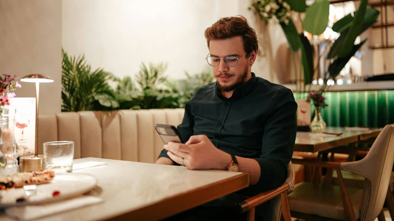 A man wearing glasses sits at a restaurant table while looking down at a smartphone.