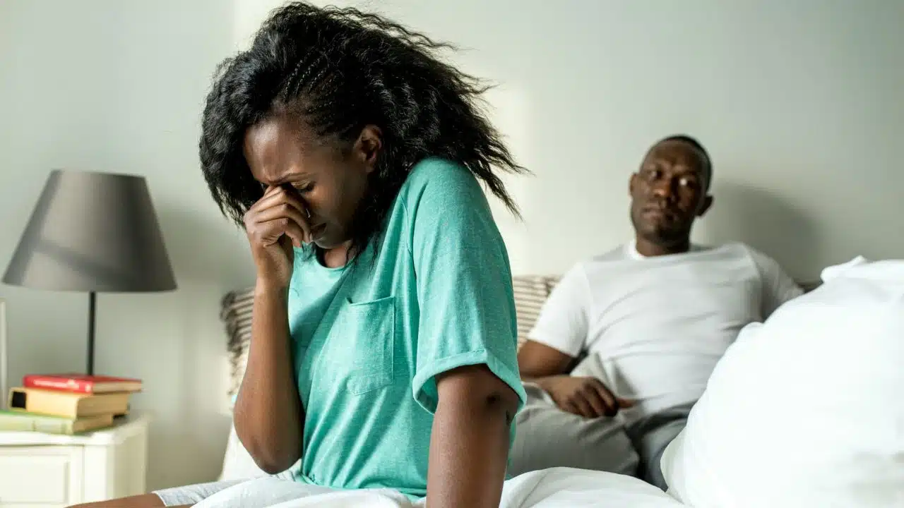 A woman sits on a bed covering her face while a man sits behind her.