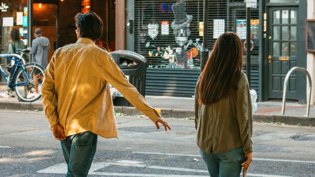 A man reaches out toward a woman walking away from him on a city street corner.